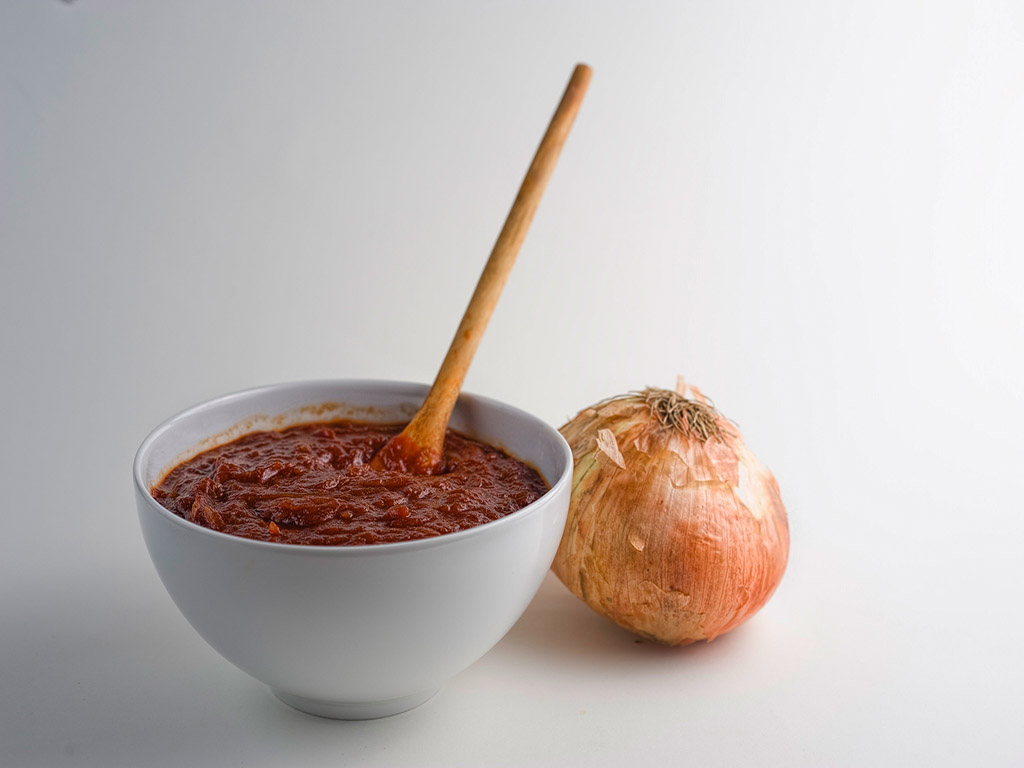 bowl of barbeque sauce with ladle inside sauce, whole onion to side of bowl on white background
